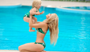 a beautiful blonde mother lifts up her happy child near an aquamarine pool